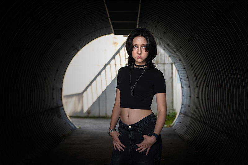 Jasmin F. (Portrait im Betontunnel) Jasmin F. steht frontal in einem runden Betontunnel mit Blick in die Kamera, gekleidet in ein schwarzes Crop-Top und dunkle Jeans.