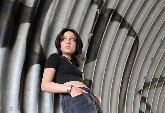 Nahaufnahme von Jasmin F., seitlich an eine gewellte Metallwand gelehnt, mit dem Blick leicht nach oben gerichtet.